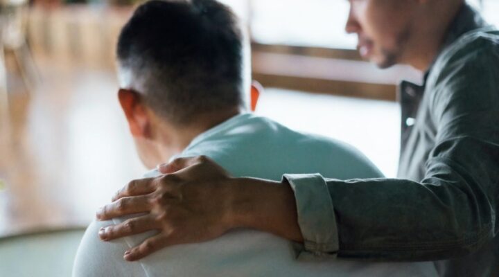 Man comforting another man by placing a supportive hand on his back, both sitting indoors with a blurred background.