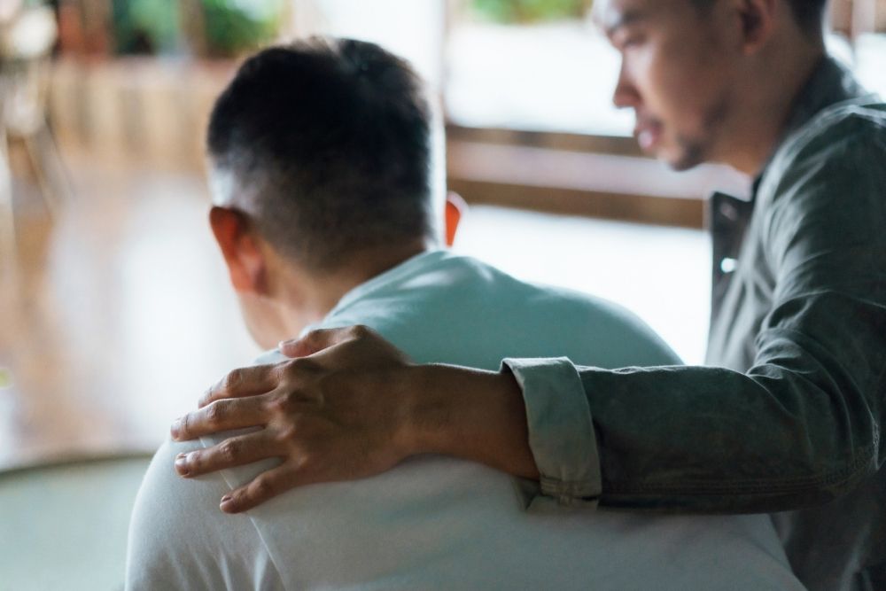 Man comforting another man by placing a supportive hand on his back, both sitting indoors with a blurred background.