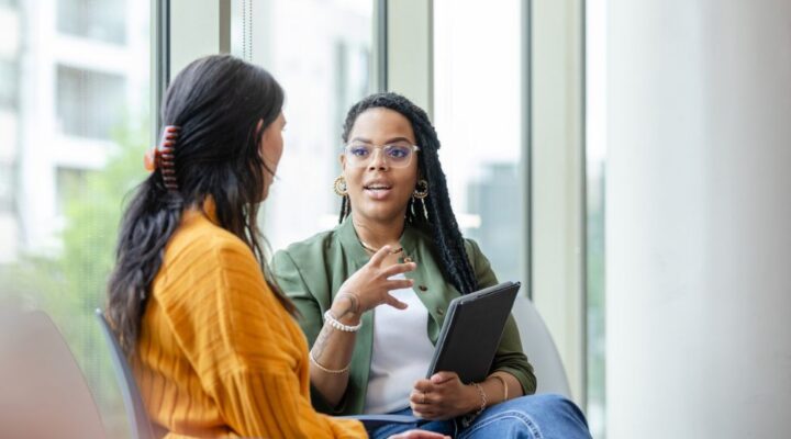 A mental health professional speaks with a client during a one-on-one therapy session. They are seated in a bright, modern office with large windows. The therapist holds a tablet while actively listening and engaging in conversation.