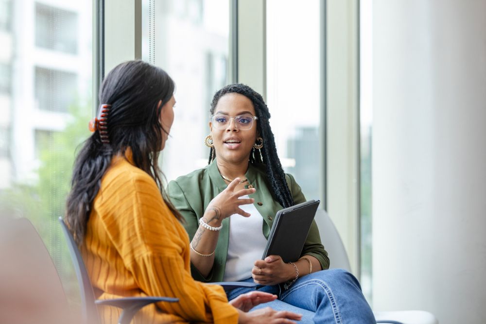 A mental health professional speaks with a client during a one-on-one therapy session. They are seated in a bright, modern office with large windows. The therapist holds a tablet while actively listening and engaging in conversation.