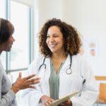 A smiling doctor holding a clipboard speaks with a patient in a bright medical office, suggesting a supportive and informative healthcare interaction.