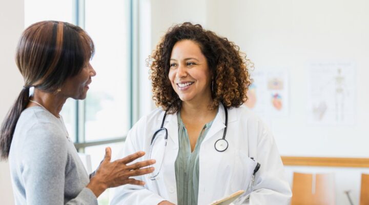A smiling doctor holding a clipboard speaks with a patient in a bright medical office, suggesting a supportive and informative healthcare interaction.