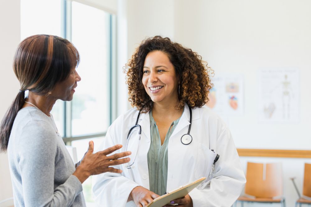 A smiling doctor holding a clipboard speaks with a patient in a bright medical office, suggesting a supportive and informative healthcare interaction.