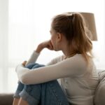 A young woman sits on a couch with her knees pulled to her chest, resting her chin on her hand. She looks out a bright window, appearing pensive or worried.