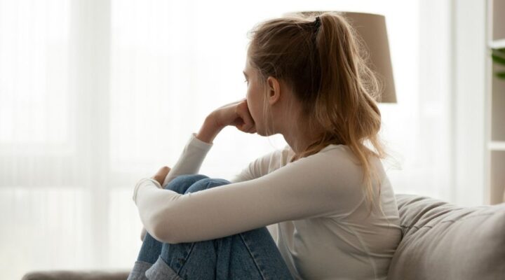 A young woman sits on a couch with her knees pulled to her chest, resting her chin on her hand. She looks out a bright window, appearing pensive or worried.