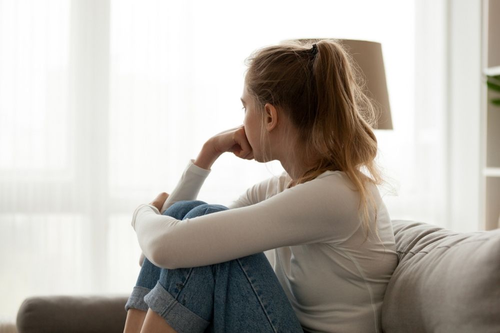 A young woman sits on a couch with her knees pulled to her chest, resting her chin on her hand. She looks out a bright window, appearing pensive or worried.