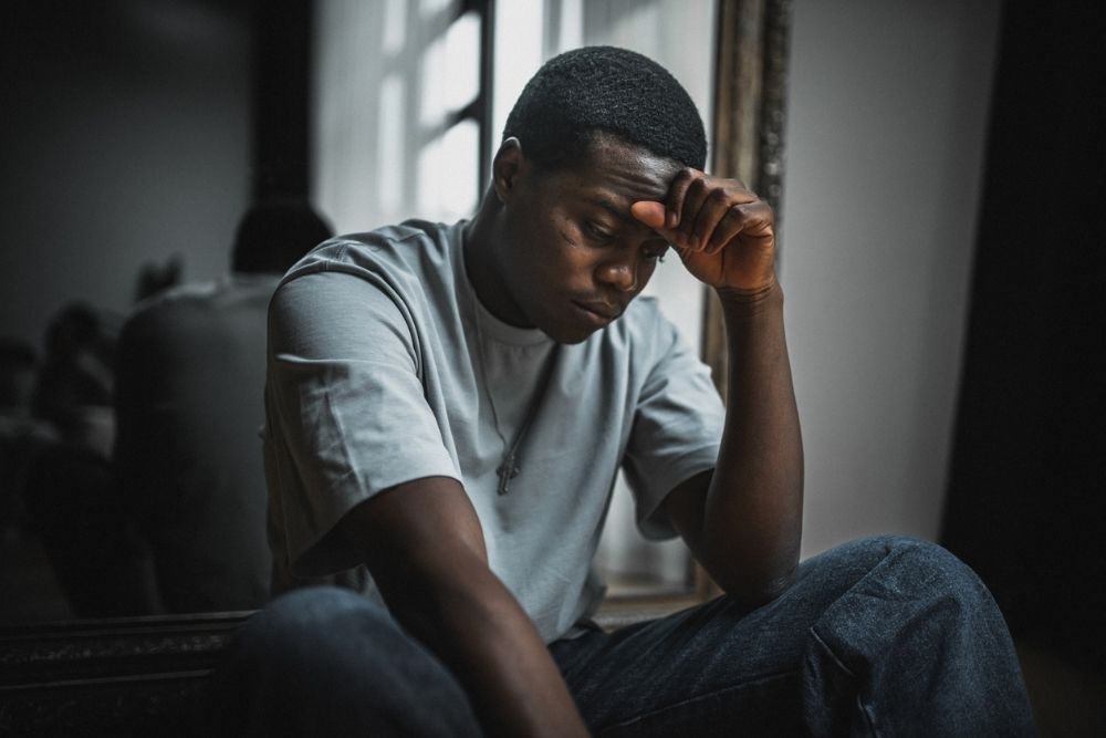 A young man sits indoors with his head resting on his hand, looking down with a pensive or distressed expression.