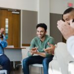 Group of men sitting in a recovery meeting, smiling and clapping in celebration, suggesting support, encouragement, and achievement of recovery milestones.