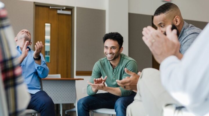 Group of men sitting in a recovery meeting, smiling and clapping in celebration, suggesting support, encouragement, and achievement of recovery milestones.
