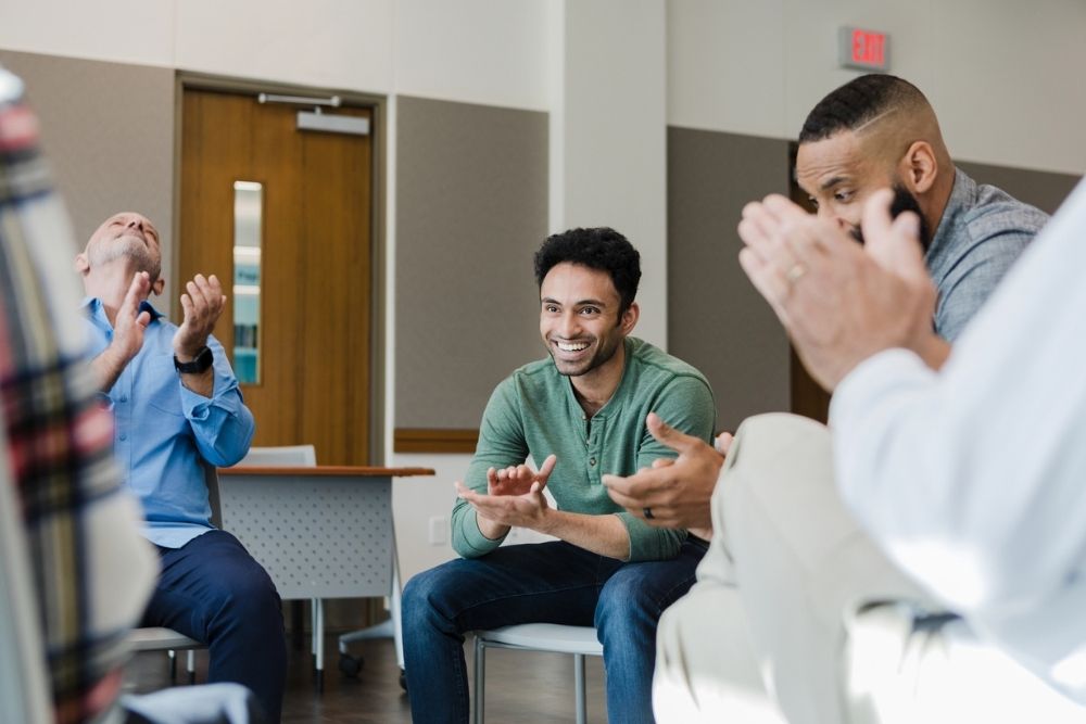 Group of men sitting in a recovery meeting, smiling and clapping in celebration, suggesting support, encouragement, and achievement of recovery milestones.