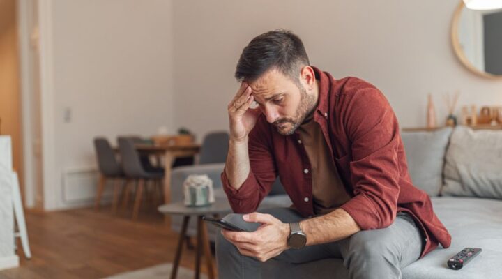 A man sitting on a couch holding his phone, looking stressed with his hand on his forehead, symbolizing struggle.