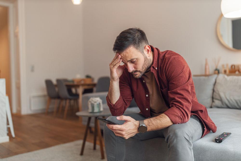 A man sitting on a couch holding his phone, looking stressed with his hand on his forehead, symbolizing struggle.