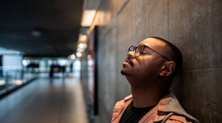 Man leaning against a wall with eyes closed, appearing stressed or deep in thought in a dimly lit hallway.