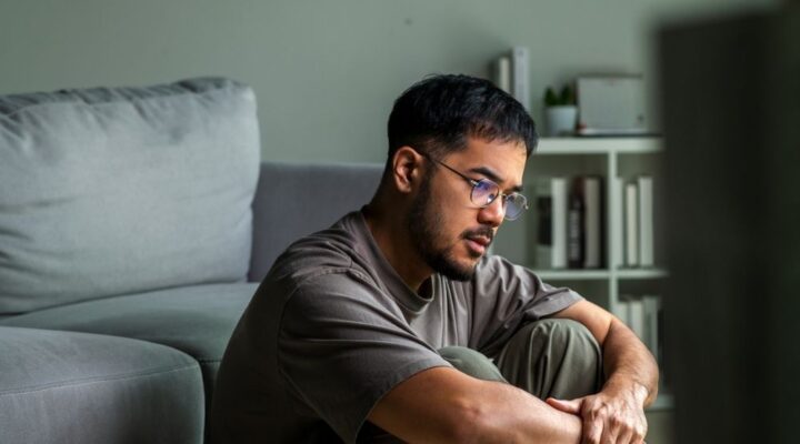 A man sits on the floor indoors with his arms wrapped around his knees, looking thoughtful and serious.