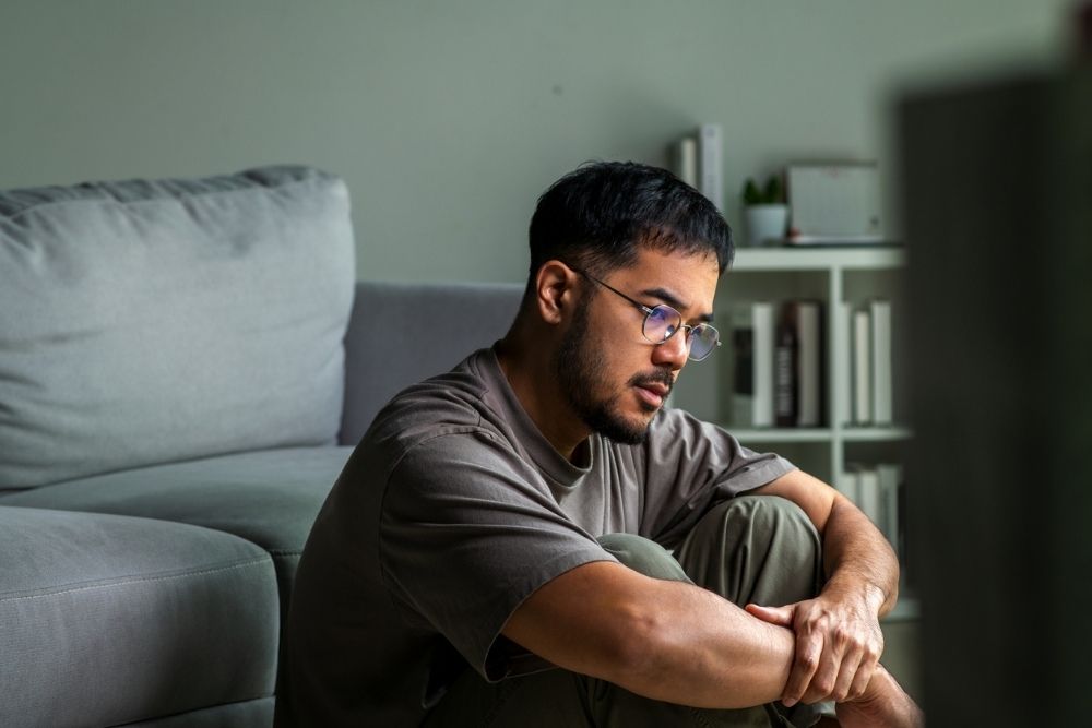 A man sits on the floor indoors with his arms wrapped around his knees, looking thoughtful and serious.