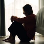 Woman sitting on the floor beside a bed with knees pulled close, appearing sad or overwhelmed in a dimly lit room.