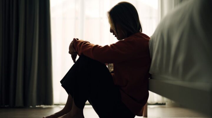 Woman sitting on the floor beside a bed with knees pulled close, appearing sad or overwhelmed in a dimly lit room.