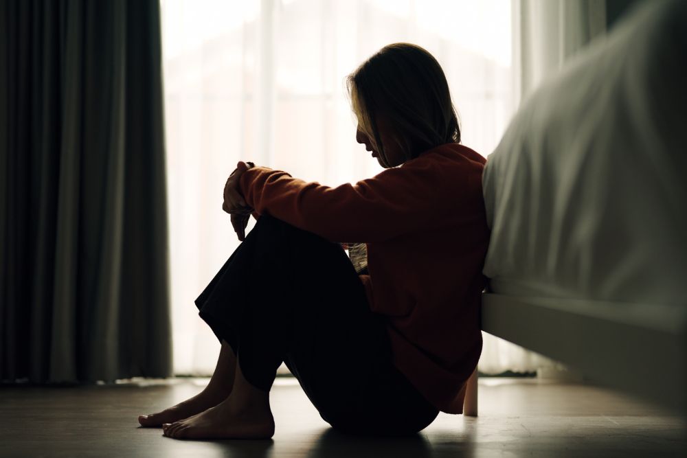 Woman sitting on the floor beside a bed with knees pulled close, appearing sad or overwhelmed in a dimly lit room.