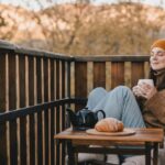 A woman sits on a wooden balcony in cool weather, wearing a beanie and cozy sweater. With her eyes closed, she holds a warm drink and relaxes next to a small table with tea and a pastry, enjoying a peaceful autumn morning.