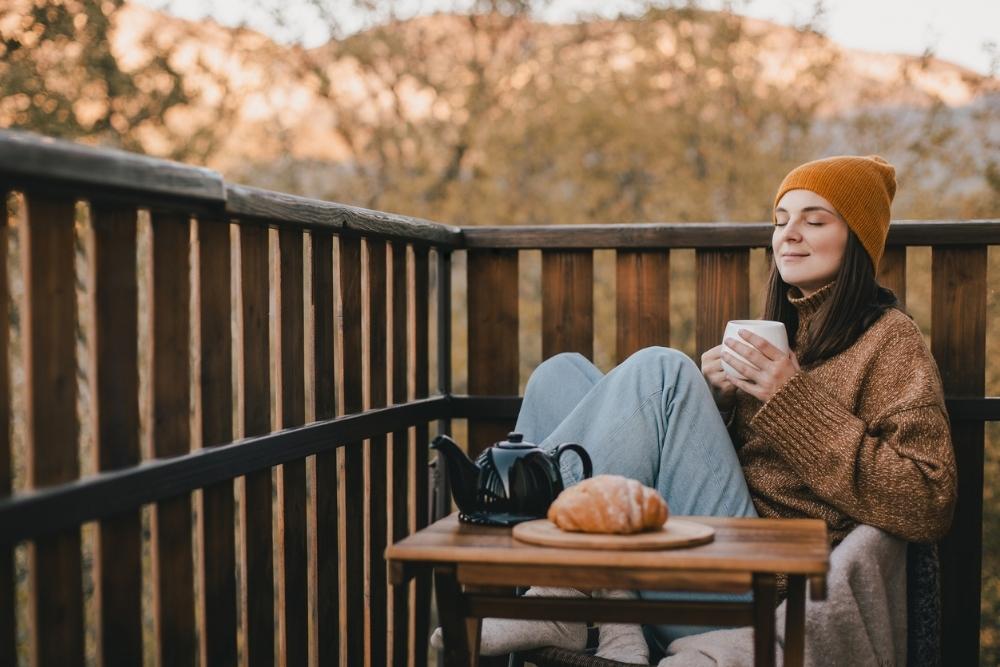 A woman sits on a wooden balcony in cool weather, wearing a beanie and cozy sweater. With her eyes closed, she holds a warm drink and relaxes next to a small table with tea and a pastry, enjoying a peaceful autumn morning.