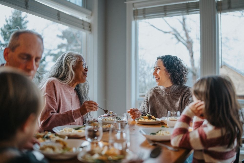 Family gathered around a dining table enjoying a meal together, with two women smiling and talking in a bright, cozy room.