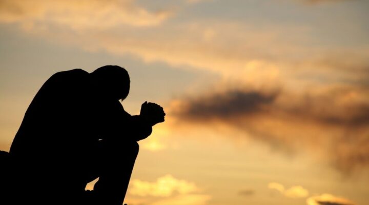 A silhouetted person sits with head bowed and hands clasped against a sunset sky, conveying reflection, struggle, or emotional distress.