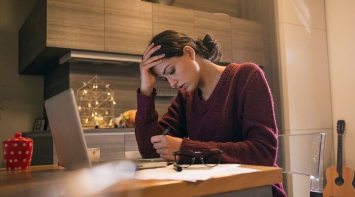 A woman sitting at a table with her head in her hand, appearing stressed while writing, with holiday decorations in the background.
