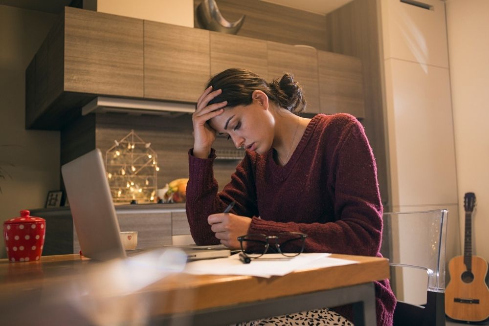 A woman sitting at a table with her head in her hand, appearing stressed while writing, with holiday decorations in the background.