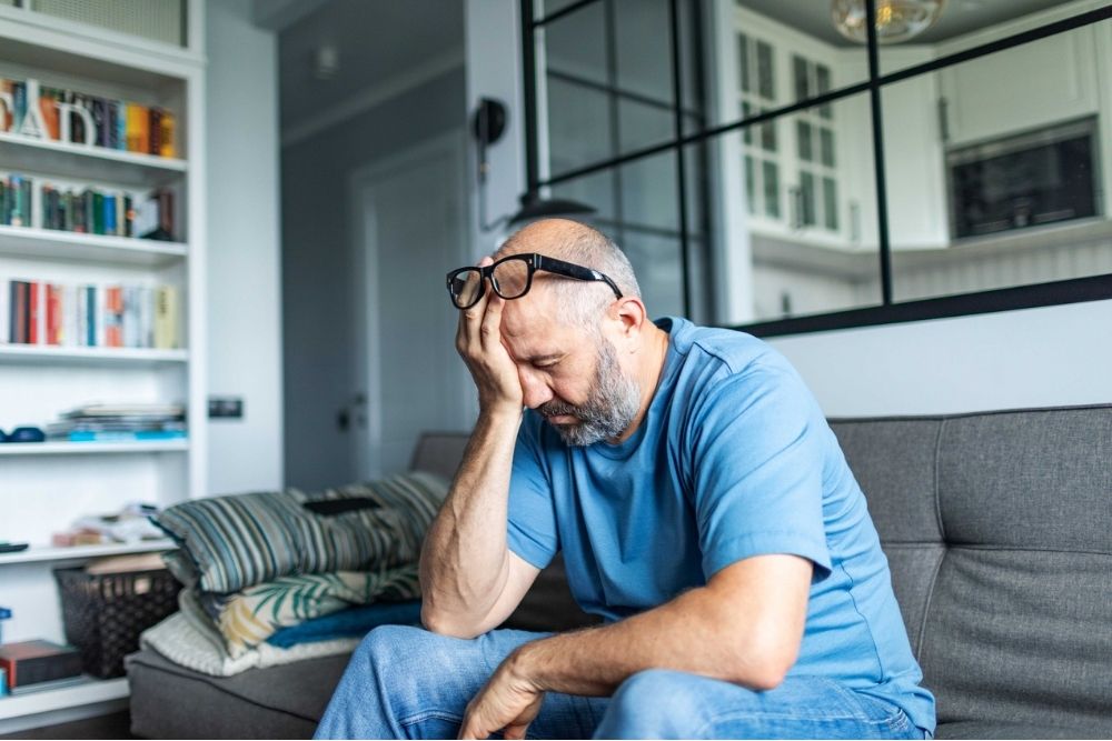 A man sitting on a couch with his head resting in his hand, appearing overwhelmed or stressed in a quiet living room.