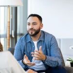A man in a blue shirt gestures while talking to a woman during a Cognitive Behavior Therapy Session. The atmosphere is calm, with natural light and a plant nearby.