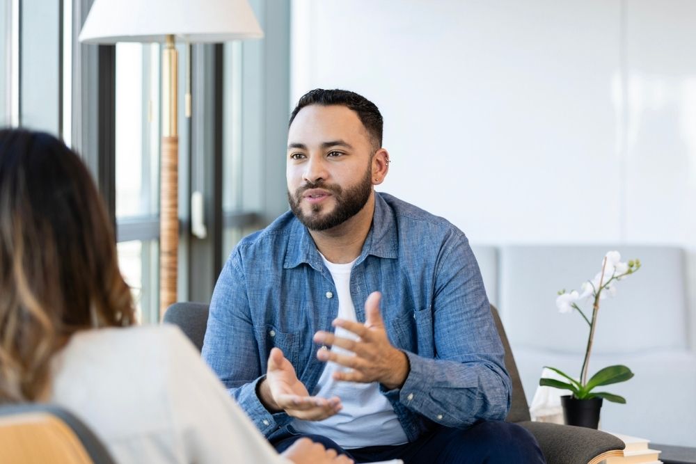 A man in a blue shirt gestures while talking to a woman during a Cognitive Behavior Therapy Session. The atmosphere is calm, with natural light and a plant nearby.