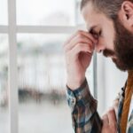 Iron Bridge - What Happens in Fentanyl Addiction Treatment. A man with a beard in a well-lit room holds his head in his hand, pinching his nose. He appears distressed.