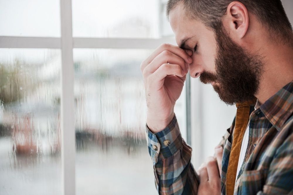 Iron Bridge - What Happens in Fentanyl Addiction Treatment. A man with a beard in a well-lit room holds his head in his hand, pinching his nose. He appears distressed.