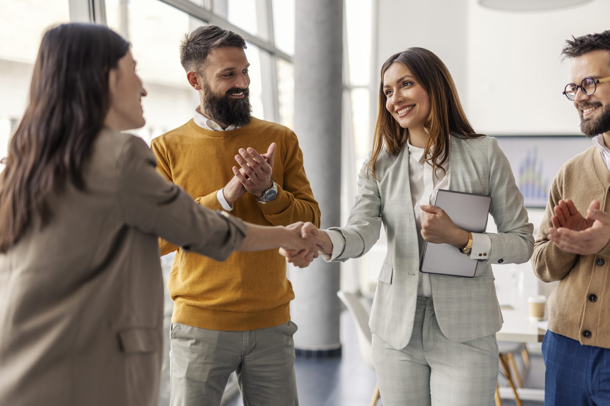 Two successful businesswomen shaking hands and closing deal and partnership while businessmen applauding them.